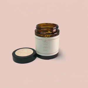 عبوة زجاجية بنية اللون مفتوحة لمنتج دخون رقم 1 من اليريا، تظهر بداخلها أقراص البخور المغلفة باللون الذهبي، وموضوعة على خلفية وردية ناعمة مع الغطاء الأسود المنفصل الذي يحمل الشعار بجانبها.