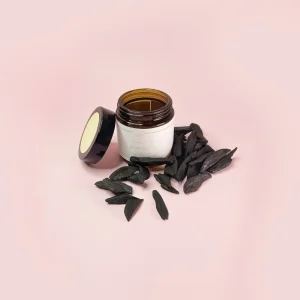 عبوة زجاجية بنية اللون مفتوحة لمنتج عود معطر 2 من اليريا، موضوعة وسط مجموعة من كسر العود الأسود المعطر الفاخر على خلفية وردية ناعمة، مع الغطاء بجانبها.